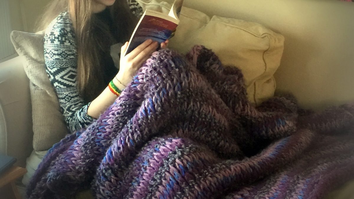 girl sitting on a couch reading a book with a purple blanket on her lap